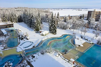 Verschneites Freibad Luftaufnahme eines verschneiten Freibads mit mehreren Becken, Dampfwasser, umgeben von Bäumen und Hotelgebäuden in einer winterlichen Landschaft.