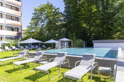 Ein Poolbereich mit Liegen, Sonnenschirmen und einem Gebäude im Hintergrund, umgeben von Bäumen.