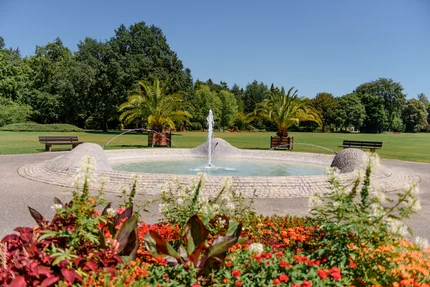 Ein runder Brunnen mit Wasserstrahl in einem Park, umgeben von Blumenbeeten und Palmen. Im Hintergrund Sicht auf Bäume und Bänke auf dem Rasen.