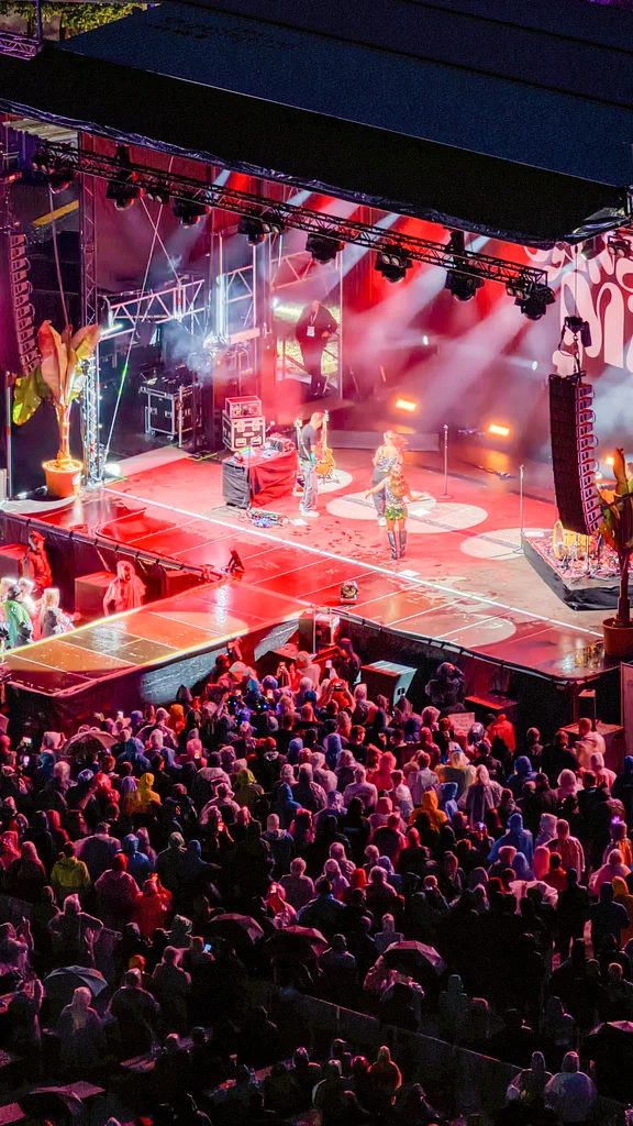 A large crowd enjoys an outdoor concert with performers on stage. The audience is mostly wearing rain gear, and the stage features colorful lighting and a large display screen.