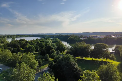 Ein Blick auf einen ruhigen Fluss umgeben von Bäumen und Wiesen unter einem blauen Himmel. Im Hintergrund sind sanfte Hügel sichtbar.