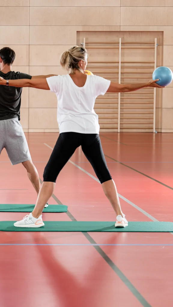 Gruppentraining in der Sporthalle Fünf Personen trainieren in einer Sporthalle und halten verschiedene Casinobälle während einer Übung. Sie stehen auf Fitnessmatten und sind in seitlicher Ausrichtung angeordnet.