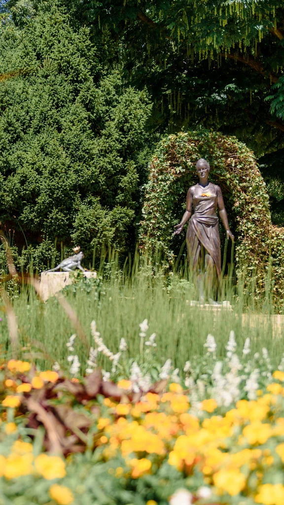 Skulptur im blühenden Garten Ein Garten mit bunten Blumen, einer Statue und einer roten Bank im Hintergrund. Grüne Büsche umrahmen die Statue, die eine Frau darstellt. Der Garten ist hell erleuchtet.