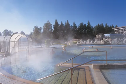 Dampfbad im Außenbecken eines Thermalbads mit mehreren badenden Personen, einer kleinen Brücke, Felsdekoration und Treppen mit Geländer.