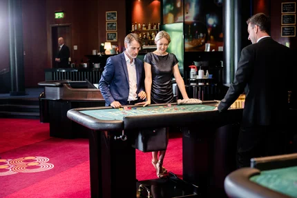 Paar am Roulette-Tisch im Casino Ein Paar steht an einem Roulette-Tisch in einem Casino. Der Mann trägt einen Anzug, die Frau ein schickes Kleid. Ein Dealer im Hintergrund ist beschäftigt. Die Szenerie ist elegant mit roten Teppichen und einer Bar im Hintergrund.