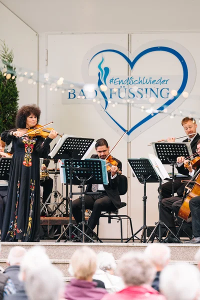 Konzert mit Orchester Eine Musikerin spielt Geige auf einer Bühne mit Orchester. Hinter ihr stehen weitere Musiker mit Instrumenten. Zuschauer sitzen davor.