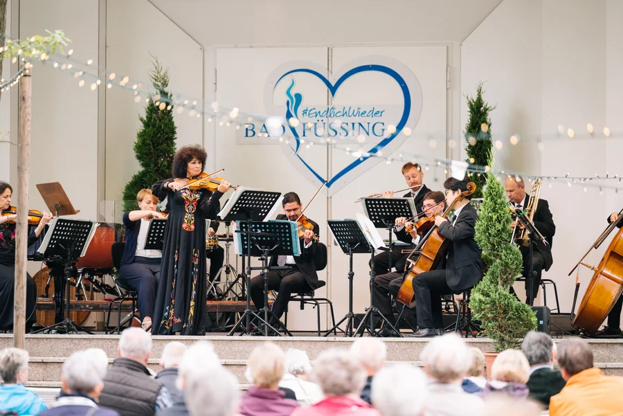 Eine Musikerin spielt Geige auf einer Bühne mit Orchester. Hinter ihr stehen weitere Musiker mit Instrumenten. Zuschauer sitzen davor.