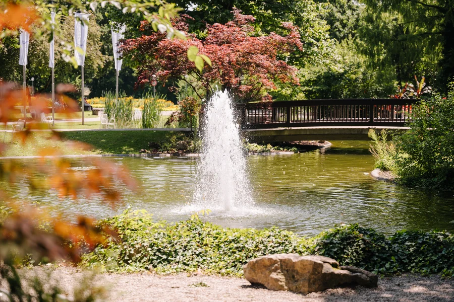 Ein Parkteich mit springendem Brunnen, kleiner Brücke, Bäumen und Blumenbeeten; im Hintergrund stehen weiße Fahnen und Stühle.