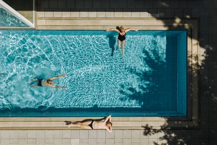 Ein Schwimmbad mit drei Personen: eine schwimmt, während zwei am Beckenrand entspannen. Das Wasser ist klar und reflektiert Licht.