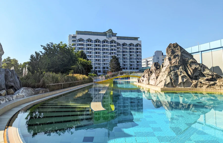 Ein großer Swimmingpool mit klarem Wasser, umgeben von Felsen und Pflanzen. Im Hintergrund steht ein großes Hotelgebäude unter klarem Himmel.