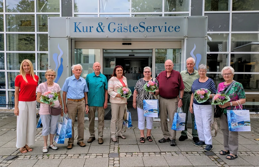 Vor einem Gebäude mit dem Schild „Kur & Gästeservice“ stehen neun ältere Menschen und eine Frau in Rot; mehrere halten Blumensträuße und Geschenktüten.