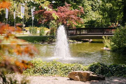 Ein stiller Park mit einem Teich, in dem ein Wasserstrahl springt. Im Hintergrund sind Bäume und eine Brücke zu sehen. Einige Sitzgelegenheiten sind um den Teich gruppiert.