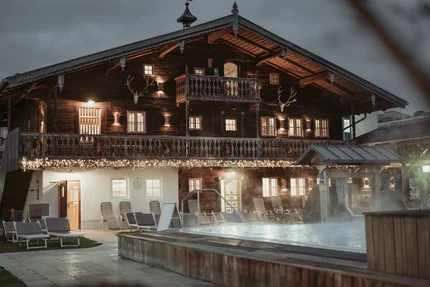 Chalet mit Außenpool Ein beleuchtetes Holzchalet bei Dämmerung mit Balkon, Hirschgeweihen an der Fassade, Liegestühlen und einem dampfenden Außenpool im Vordergrund.