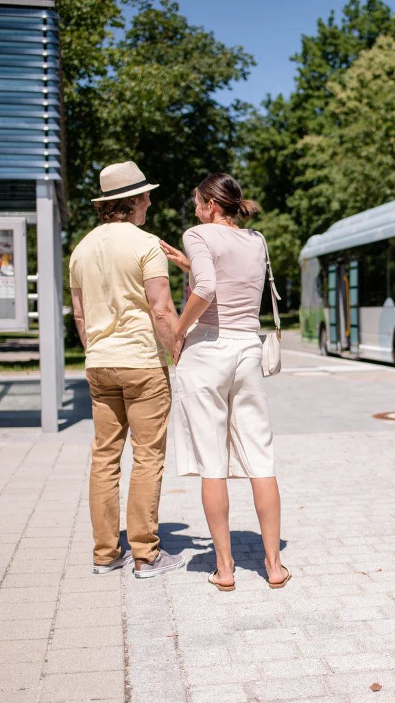 Ein Paar steht an einer Bushaltestelle und hält Händchen, während ein Bus in der Nähe wartet. Der Hintergrund zeigt Bäume und Blumen.