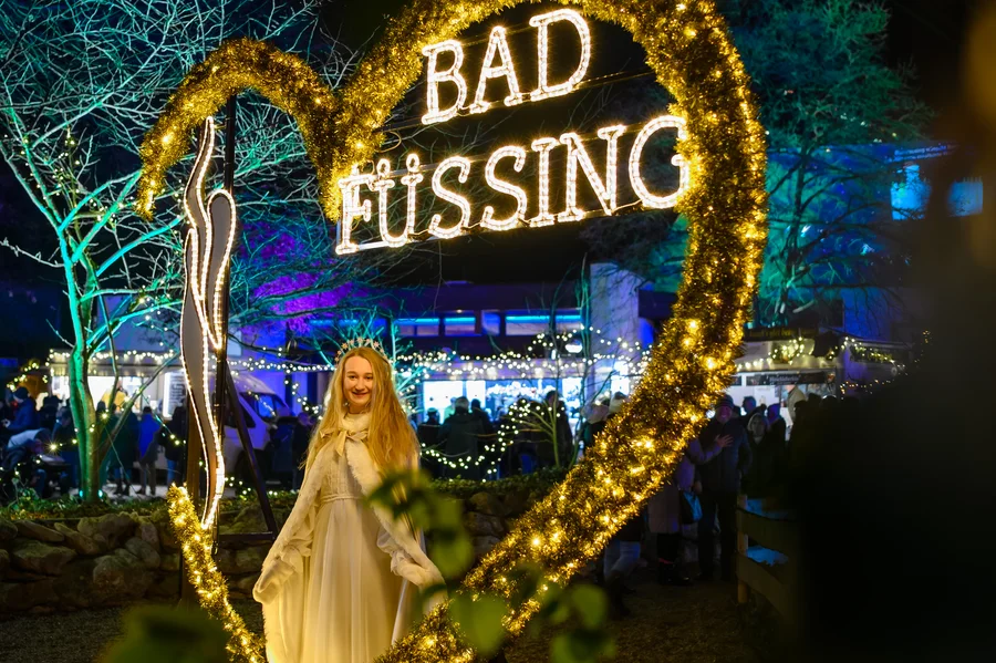 Frau vor Lichtdekoration in Bad Füssing Eine Frau in einem weißen Kleid steht vor einem hell erleuchteten Herz mit den Worten 'BAD FÜSSING'. Die Szenerie ist festlich mit bunten Lichtern im Hintergrund.
