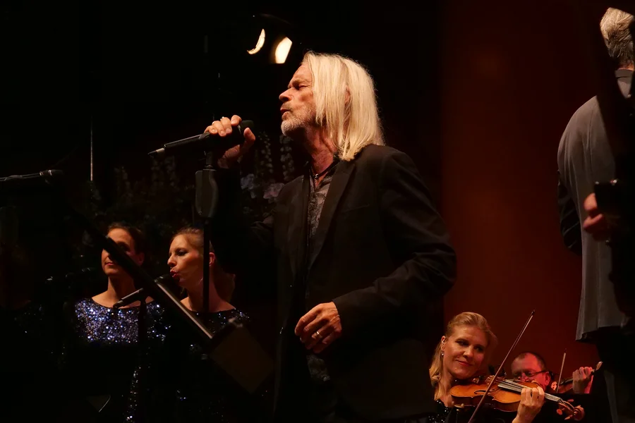 Ein Sänger mit langen, grauen Haaren steht mit einem Mikrofon auf der Bühne. Im Hintergrund sind Frauen in glitzernden Kleidern und Musiker zu sehen. Eine Violinistin lächelt, während sie auf ihrem Stuhl sitzt.