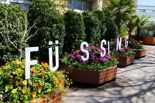Bunte Blumen mit dem Wort Füßing Ein bepflanzter Bereich mit bunten Blumen und großen Buchstaben, die das Wort ‚Füßing‘ bilden. Umgeben von grünen Sträuchern und Bäumen.