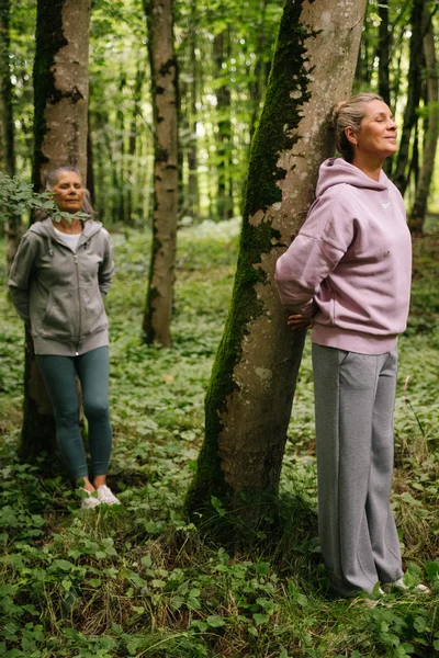 Drei Frauen stehen in einem Wald, lehnen sich an Bäume. Sie wirken entspannt und meditativ, umgeben von grüner Vegetation.