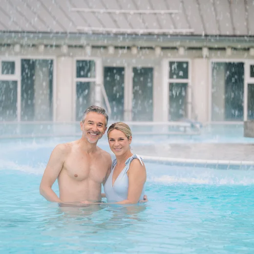 Ein Mann und eine Frau stehen in einem Pool mit Wasserfällen im Hintergrund. Der Mann lächelt, hat graue Haare und ist oben ohne. Die Frau trägt einen hellblauen Badeanzug und lächelt ebenfalls. Im Hintergrund sieht man Fenster und einen Wasserfall.