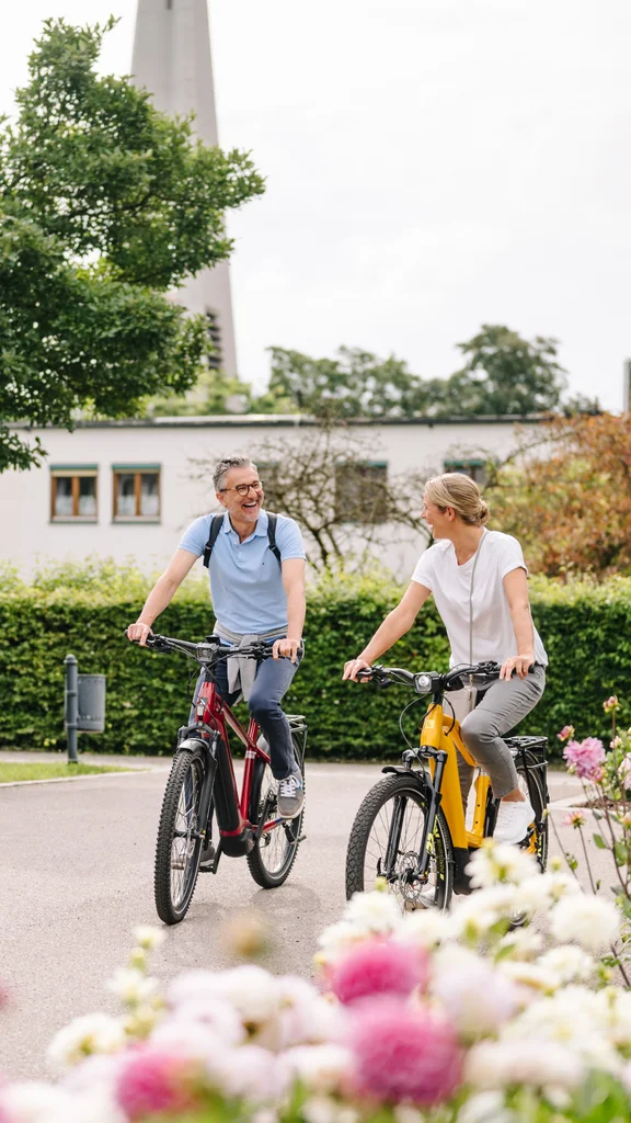 Ein Mann und eine Frau fahren auf E-Mountainbikes in einem Garten mit Blumen. Im Hintergrund sind Bäume und ein Gebäude sichtbar.