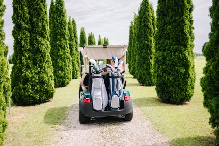 Golfwagen zwischen Lebensbäumen Ein Golfwagen mit Golftaschen fährt auf einem Schotterweg, umgeben von hohen Zypressen. Im Hintergrund ist eine Wiese sichtbar.