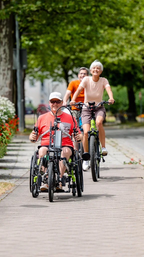 Eine Person in einem Handbike und zwei Radfahrer, die auf einem Bürgersteig entlangfahren, umgeben von grünen Bäumen und blühenden Blumen.