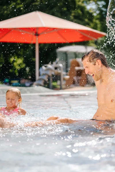 Ein Vater spielt mit seiner Tochter im Wasser. Der Vater sitzt im Pool und lacht, während die Tochter im Wasser spritzt. Ein rotes Sonnenschirm ist im Hintergrund sichtbar.