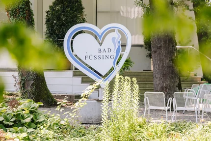 Bad Füssing Schild Ein herzförmiges Schild mit der Aufschrift "Bad Füssing" steht vor einer gepflegten Gartenlandschaft. Grüne Pflanzen und Stühle sind im Hintergrund sichtbar.