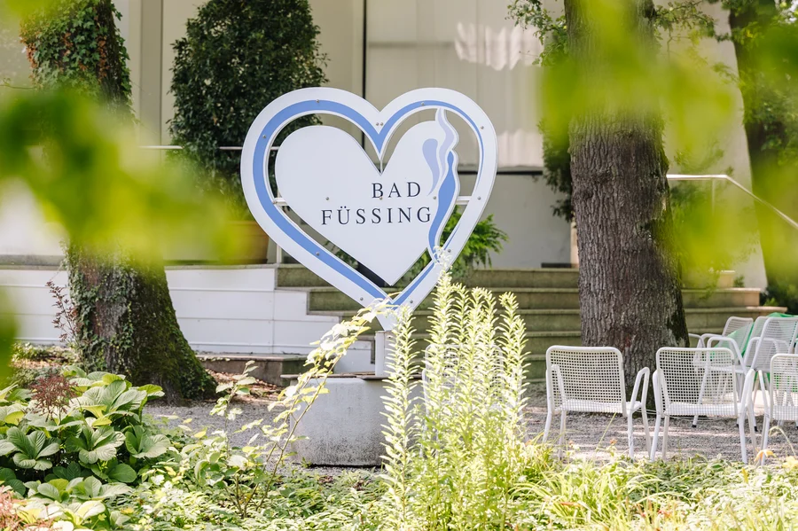 Bad Füssing Schild Ein herzförmiges Schild mit der Aufschrift "Bad Füssing" steht vor einer gepflegten Gartenlandschaft. Grüne Pflanzen und Stühle sind im Hintergrund sichtbar.