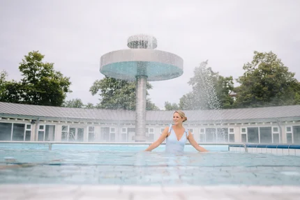 Eine Frau steht in einem Pool mit einem Wasserfall im Hintergrund. Der Pool hat eine blaue Farbe und die Umgebung ist grün mit Bäumen. Der Himmel ist bewölkt.