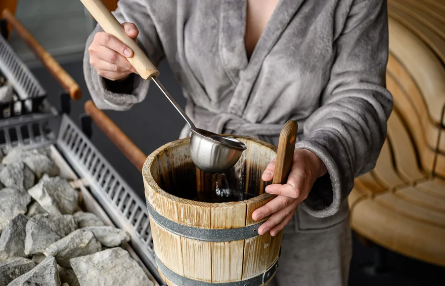 Person schöpft Wasser aus Eimer Eine Person in einem Bademantel schöpft mit einem Löffel Wasser aus einem Eimer, der neben einem Saunasteinbereich steht.