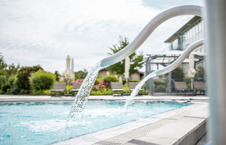 Ein Pool mit Wasserfontänen und Gartenlandschaft im Hintergrund. Die Sonne scheint, und es sind Liegen und Schirme sichtbar.