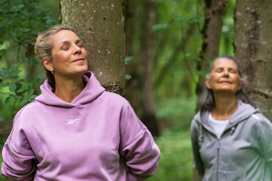 Zwei Frauen entspannen in einem Wald, eine lehnt an einem Baum. Beide haben geschlossene Augen und lächeln, während sie die Natur genießen.