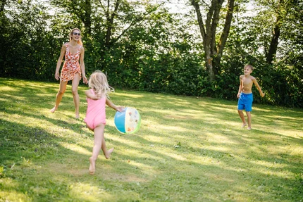 Eine Frau und zwei Kinder spielen im Freien mit einem Ball. Die Frau trägt ein buntes Kleid und die Kinder sind in Schwimmbekleidung. Der erste Junge ist ohne Shirt, der zweite in einem rosa Badeanzug. Im Hintergrund sind Bäume zu sehen.