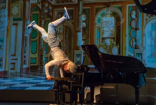 Akrobat auf Klavier bei Performance Eine Person steht auf den Händen, während sie auf einem Klavier sitzt. Im Hintergrund ist ein bunter Raum mit historischen Elementen zu sehen.