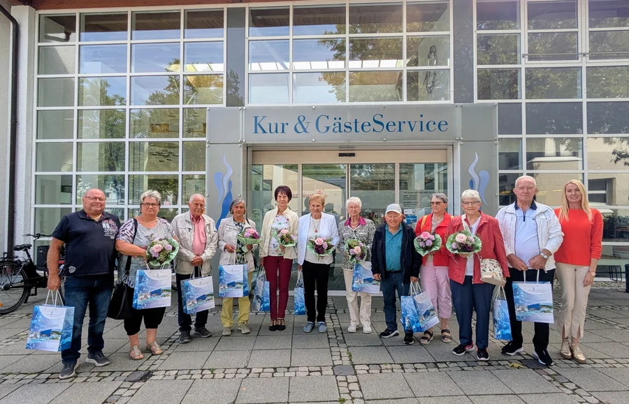 Eine Gruppe von älteren und jüngeren Menschen steht vor dem Eingang von „Kur & GästeService“ und hält Blumensträuße sowie Geschenktüten in den Händen.