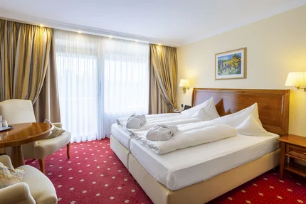 Ein gemütliches Hotelzimmer mit Doppelbett, zwei Kissen und frischer Bettwäsche. Ein kleiner Tisch mit zwei Stühlen steht auf einem blumigen roten Teppich. Vorhänge verdecken Fenster mit Tageslicht und ein Gemälde hängt an der Wand.