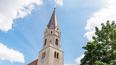 Hoher Kirchturm bei blauem Himmel Der hohe Kirchturm ist aus grauem Stein und hat ein spitzes Dach mit einem Kreuz. Einige Fenster sind sichtbar, und der Himmel ist blau mit Wolken.