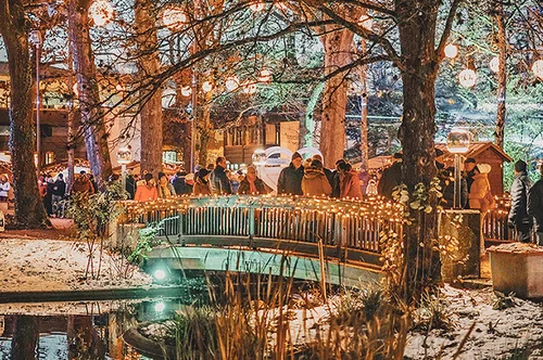 Winterliche Beleuchtung im Park Eine winterliche Szene mit festlich beleuchteten Bäumen, einem beleuchteten Brückenbogen über einen ruhigen Teich und einer Menschenmenge, die sich in der Nähe versammelt. Schnee ist am Boden sichtbar.