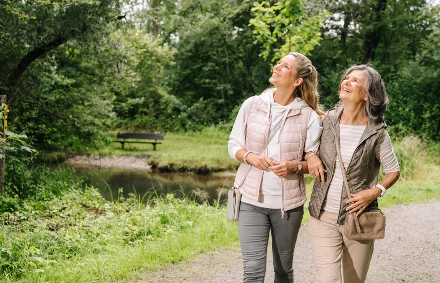 Zwei Frauen gehen nebeneinander auf einem Weg durch einen grünen Park, lächelnd und gestikulieren, während sie nach oben schauen. Im Hintergrund ist ein ruhiger Teich und eine Bank zu sehen.
