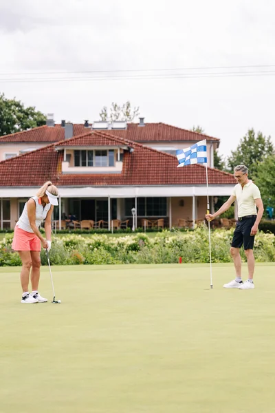 Eine Frau puttet auf dem Golfplatz, während ein Mann die Fahne hält. Im Hintergrund befindet sich ein Gebäude mit rotem Dach und grünen Bäumen umgeben.