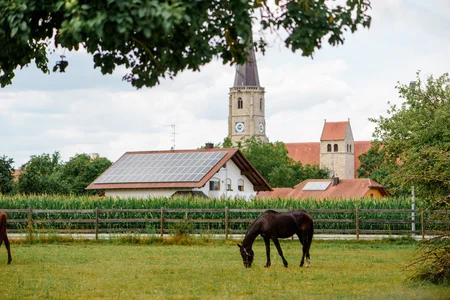 Ländliche Szene mit Pferd Ein schwarzes Pferd grast auf einer Wiese vor einem Dorf mit zwei Kirchen und einem Haus mit Solarpanelen. Im Hintergrund sind Bäume und ein Maisfeld zu sehen.