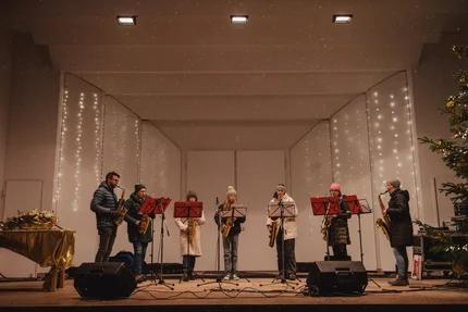 Weihnachtskonzert mit Saxophonisten Eine siebenköpfige Band spielt auf einer Bühne. Mitglieder spielen Saxophon und tragen warme Kleidung. Der Hintergrund ist mit Lichtern dekoriert.