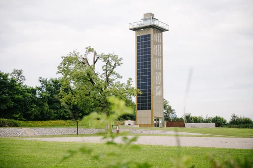 Ein moderner Aussichtsturm aus Glas und Metall steht in einer grünen Landschaft mit Bäumen und Wiese. Der Himmel ist bewölkt und die Umgebung wirkt friedlich.