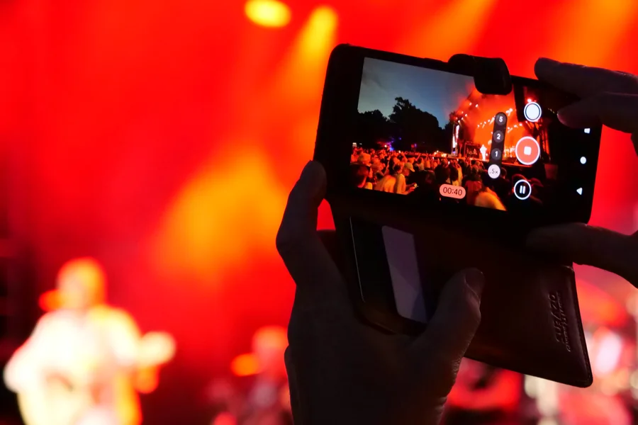 A person holding a smartphone recording a concert with bright orange lights in the background and a blurred musician in the foreground.
