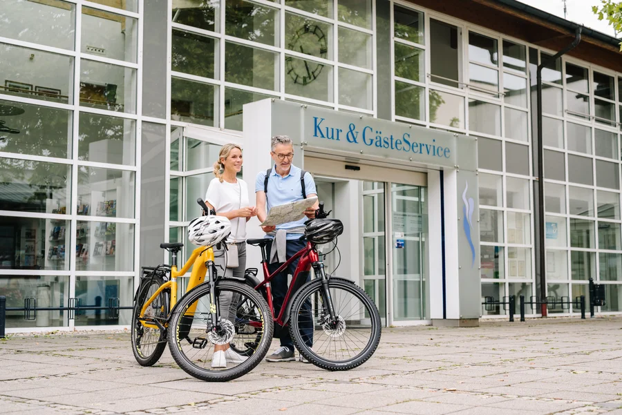 Ein Mann und eine Frau stehen vor einem Gebäude mit einem Schild "Kur & GästeService". Der Mann hält eine Landkarte, während die Frau interessiert zuschaut. Neben ihnen stehen zwei Fahrräder, eines in gelb und eines in rot.