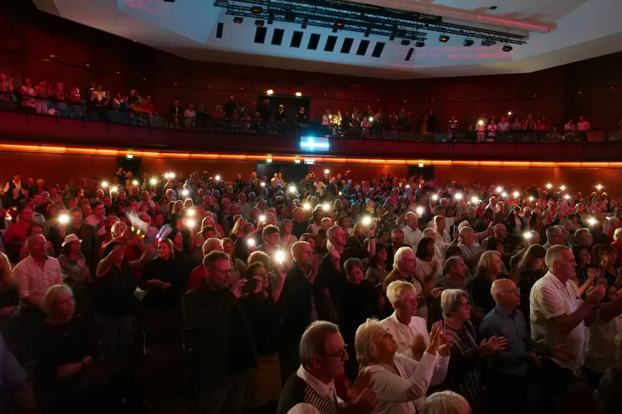 Eine große Menschenmenge in einem Veranstaltungsort, die applaudiert und ihre Handys mit Licht zeigt. Viele Menschen stehen mit Freude, während einige oben im Balkonbereich stehen.
