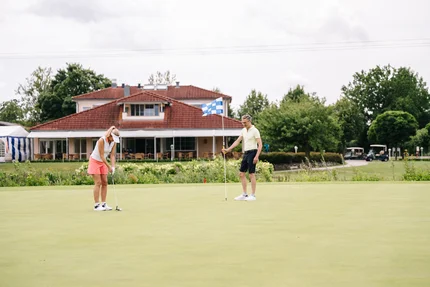 Golfspiel auf einem Putting Green Eine Frau mit einem Golfball bereit zum Putten. Ein Mann steht am Golfloch mit einer Flagge. Im Hintergrund ein Golfclubhaus mit Tischen und Stühlen.