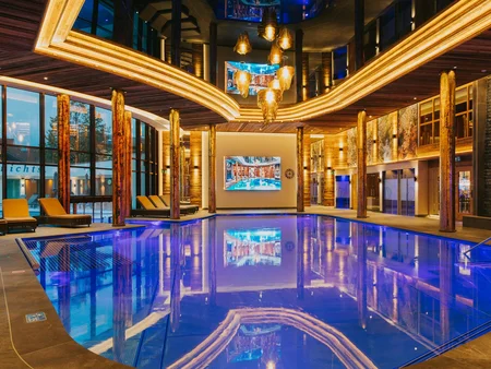 Innenpool mit blauen Lichtern Ein moderner Innenpool mit blauen Lichtern und großzügigen Fenstern. Sonnenliegen stehen am Rand, und eine große Leinwand zeigt ein Bild. Holzelemente verleihen dem Raum eine natürliche Atmosphäre.