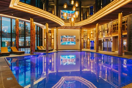 Innenpool mit blauen Lichtern Ein moderner Innenpool mit blauen Lichtern und großzügigen Fenstern. Sonnenliegen stehen am Rand, und eine große Leinwand zeigt ein Bild. Holzelemente verleihen dem Raum eine natürliche Atmosphäre.